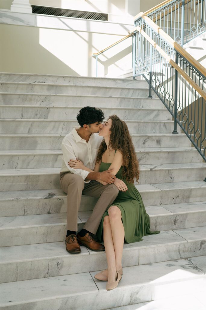 couple sitting on marble stairs at a library kissing 