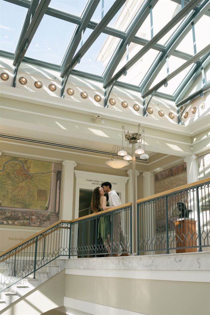 woman leaning against rail while her boyfriend is kissing her in a library with a ton of windows 