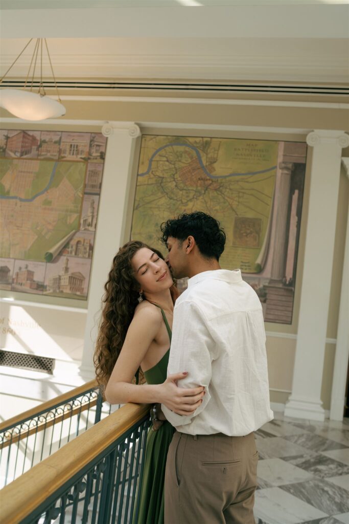 man kissing his partner on the cheek while she is leaning against a railing 